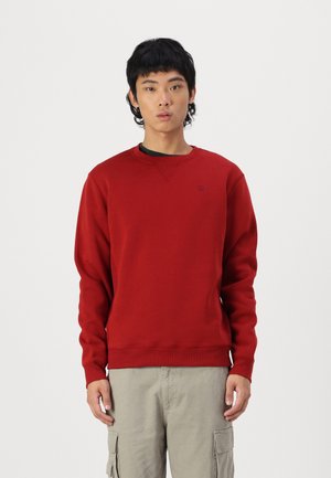 Felpa rossa con scollo rotondo, maniche lunghe e polsini a costine. Presenta un piccolo logo sul petto e una texture morbida. Indossata sopra pantaloni cargo chiari.