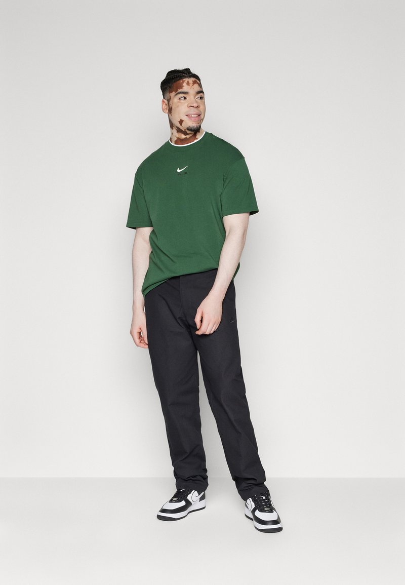 T-shirt en coton vert avec un petit logo Nike blanc, associé à un pantalon noir et des baskets noir et blanc. Le modèle se tient contre un fond uni.