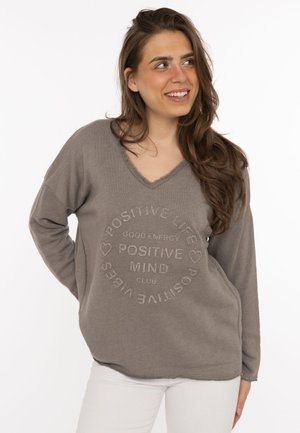 Lächelnde Frau mit langen braunen Haaren, die einen taubenfarbenen V-Ausschnitt-Pullover mit dem Schriftzug "Positive Life, Positive Mind, Positive Vibes" und weiße Hosen trägt.