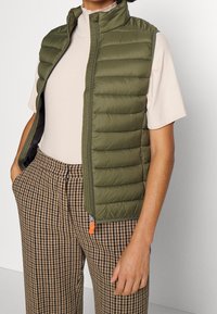 Oliwkowa kamizelka puffer z wysokim kołnierzem, zamkiem błyskawicznym i żebrową fakturą. Stylizowana na beżowej bluzce i spodniach w kratę houndstooth.