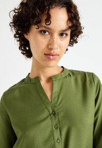 Chemise verte à col mao, fabriquée en tissu lisse. Elle présente une patte de boutonnage avec cinq boutons et des détails de couture minimalistes.