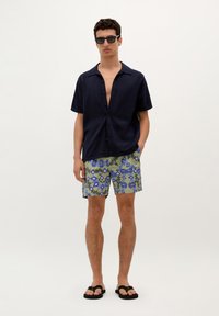Chemise à manches courtes navy avec boutons, short vert à motifs avec des formes bleues, et sandales noires, offrant une coupe décontractée et un design informel.