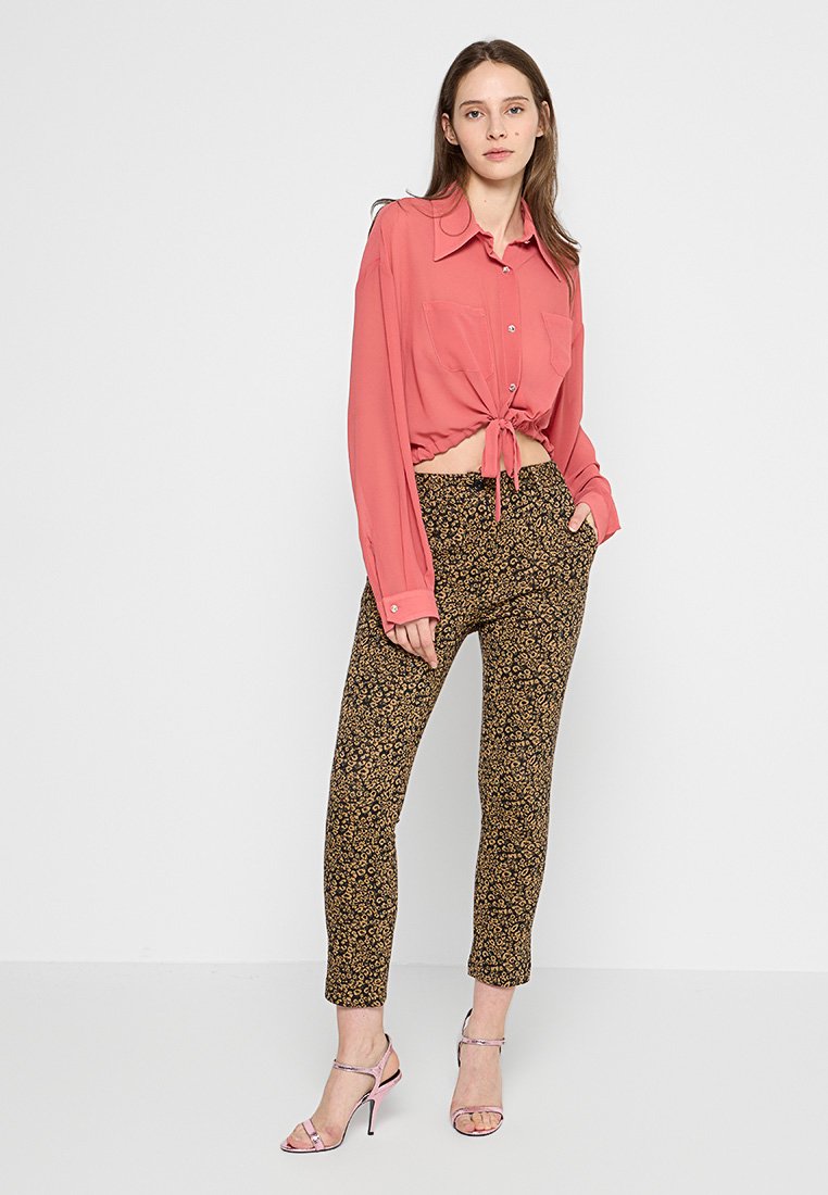 Rosa langärmlige Bluse mit Knoten vorne, kombiniert mit braunen Hose mit Blumenmuster. Das Model trägt silberne Riemchen-Heels. Einheitlicher weißer Hintergrund.