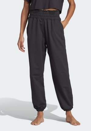 Osoba ubrana w czarne spodnie dresowe z elastycznym pasem oraz czarny krótki top, stojąca boso na neutralnym tle.