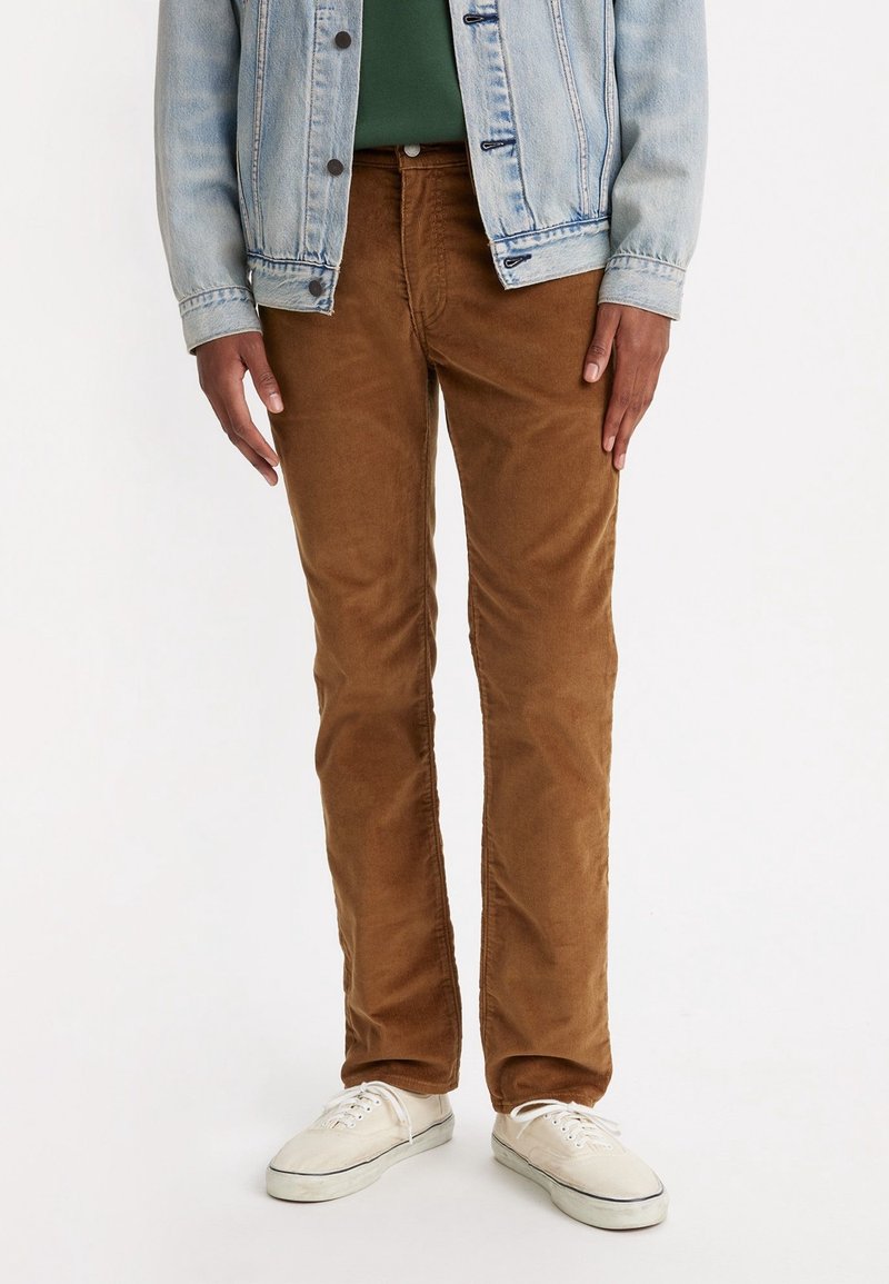 Homme portant un pantalon en velours marron, des baskets blanches, une chemise verte et une veste en jean bleu clair, debout sur un fond blanc.