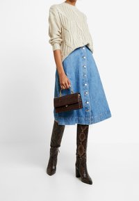 Personne portant un pull en maille torsadée beige, une jupe midi en jean bleu avec boutons sur le devant, des bottes hautes en peau de serpent jusqu'aux genoux, tenant un sac à main marron foncé avec une poignée dorée.