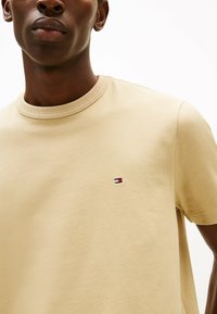 Beżowa koszulka typu crew neck z miękkiej bawełny, z małym haftowanym logo w kolorze czerwonym, białym i niebieskim na lewej piersi. Gładka tekstura.