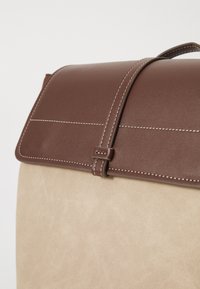 Flap en cuir brun avec une fermeture à pression au-dessus d'un corps en daim beige doux. Des coutures blanches mettent en valeur le design. Forme minimaliste et structurée.