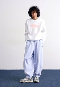 Sweatshirt branco da Nike com logótipo vermelho, combinado com calças de treino azul claras. A pessoa usa ténis prateados com solas brancas. Cabelo encaracolado.