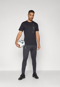 Svart trenings t-skjorte med logo og tekstdetaljer, kombinert med grå tights som sitter tight med logo. Holder en mønstret fotball.
