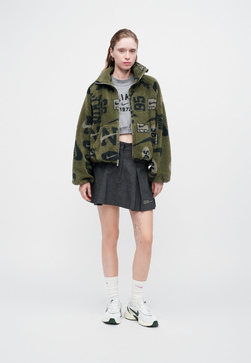 Fleecejacke in olivgrün mit schwarzen Mustern, graues kurz geschnittenes T-Shirt, grauer Faltenrock, weiße Socken und weiße/grüne Turnschuhe.