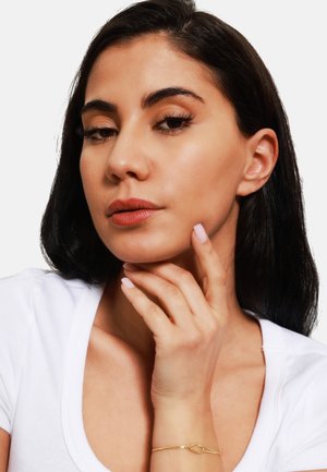 Nahaufnahme einer Person mit mittlerem Hautton, dunklem Haar, die ein weißes Oberteil trägt. Goldarmband am Handgelenk, hellrosa Nagellack, dezentes Makeup.