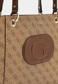Brun tekstureret shoppertaske med en fremtrædende præget "G"-logo og et guldfarvet metalaccent med teksten "GUESS" på forsiden.