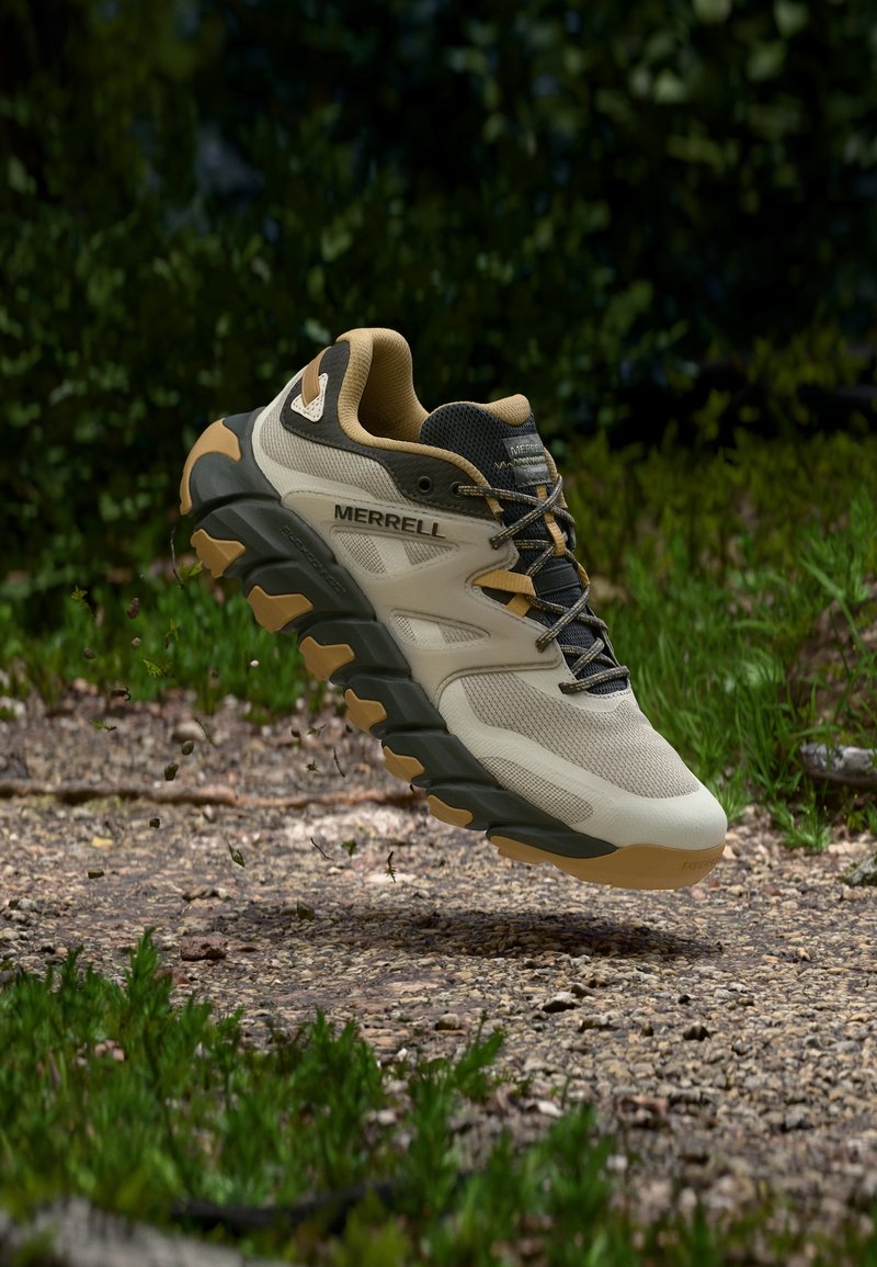 Ελαφρώς καφέ και μαύρο πεζοπορικό παπούτσι Merrell στον αέρα πάνω από βραχώδη μονοπάτι περιτριγυρισμένο από πράσινα φυτά και με θαμπό φόντο το δάσος.