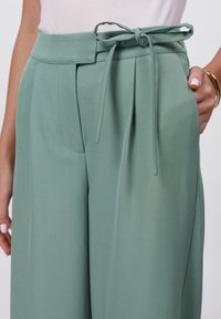 Wide-leg trousers in textured mint fabric, featuring a tie belt at the waist, front pleats, and side pockets. Smooth finish.