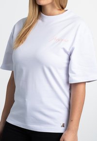 White cotton t-shirt with short sleeves, featuring a light pink embroidered logo on the chest and a metallic accent detail on the hem.