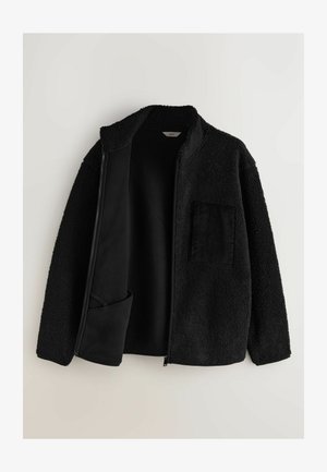 Czarna kurtka zapinana na zamek, wykonana z teksturowanej, puszystej tkaniny, z miękkim wnętrzem. Posiada kieszeń z kordurowego materiału po lewej stronie.