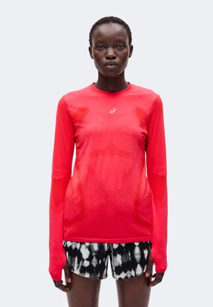 Femme portant un t-shirt de sport à manches longues rouge vif et un short à motifs noir et blanc, debout face à l'avant sur un fond uni.