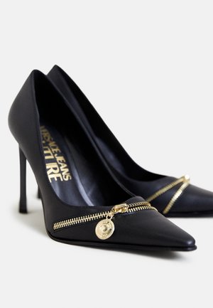 Sorte højhælede sko med spids tå, med en dekorativ gylden lynlås og vedhæng foran. Glat lædertekstur.