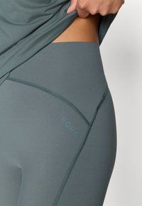 Szürke, magas derekú leggings sima, rugalmas anyagból, derék körüli logóval és kontrasztos varrásokkal.