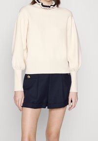 Cremesweater mit puffärmligen Ärmeln und einem schwarzen Rüschkragen, kombiniert mit dunkelblauen Shorts mit goldenen Knopfakzenten und seitlichen Taschen.
