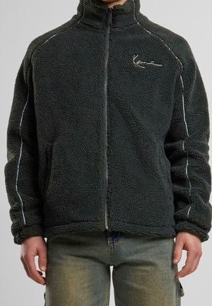 Zwarte fleecejas met rits, witte biezen op de mouwen en een logo op de borst, gedragen over verweerde blauwe spijkerbroek.