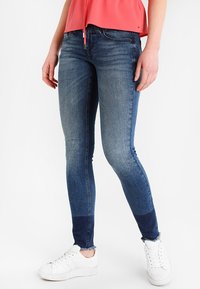 Jeans ajustados en denim azul medio con una textura desteñida. Presentan paneles de un azul más oscuro en el dobladillo. Combinados con zapatillas blancas.
