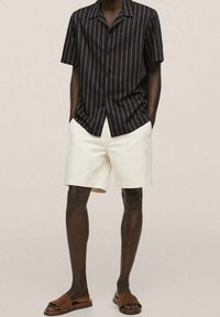 Uomo che indossa una camicia nera a maniche corte con bottoni e sottili righe rosse, pantaloni corti bianchi e sandali marroni, in piedi con le mani in tasca.