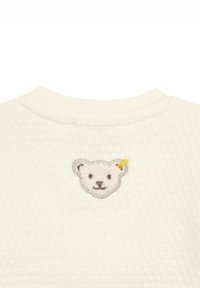 Sweatshirt beige texturé avec un col rond. Il présente une applique de visage d'ours cousue avec un accent jaune à l'arrière.