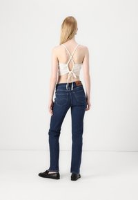 Top corto blanco con tirantes y lazo en la parte de atrás, combinado con jeans ajustados de color azul oscuro que presentan un parche de cuero con el logo y bolsillos traseros clásicos.