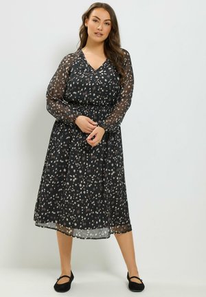 Vrouw met lang haar, gekleed in een zwarte doorschijnende midi-jurk met lange mouwen en een gespikkeld patroon, en zwarte platte schoenen, staand tegen een witte achtergrond.
