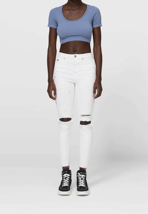 Femme portant un haut court bleu à manches courtes, un jean skinny blanc déchiré et des baskets montantes noires, debout devant un fond uni.
