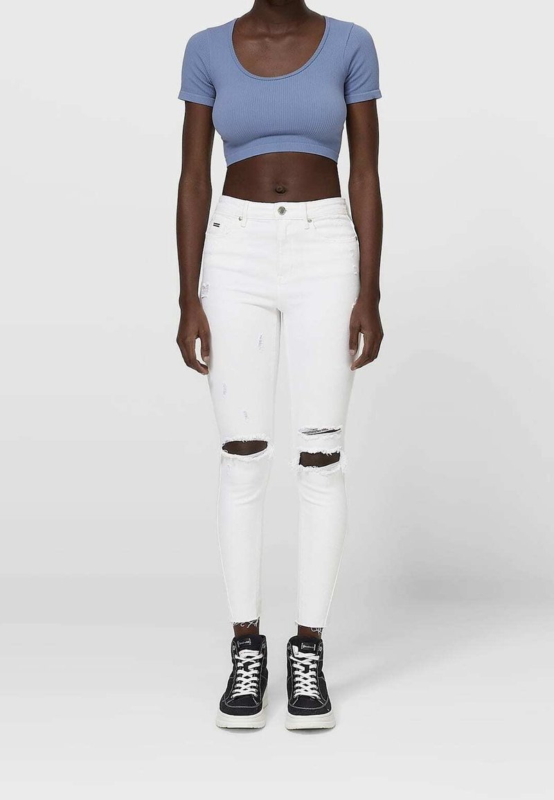 Femme portant un haut court bleu à manches courtes, un jean skinny blanc déchiré et des baskets montantes noires, debout devant un fond uni.