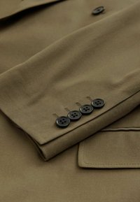 Brūns teksturēts audums ar piedurknēm, kurām ir četri melni pogas un redzamie šuvju detaļas, demonstrējot pielāgotu dizainu.