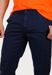 Navy blue cotton pants featuring a button closure, front pockets, and a smooth texture. Worn with an orange short-sleeve shirt.