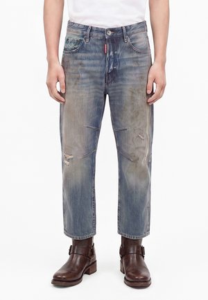 Homme portant un jean bleu délavé et usé avec des coutures aux genoux et des bottines en cuir marron, debout les mains détendues le long du corps.