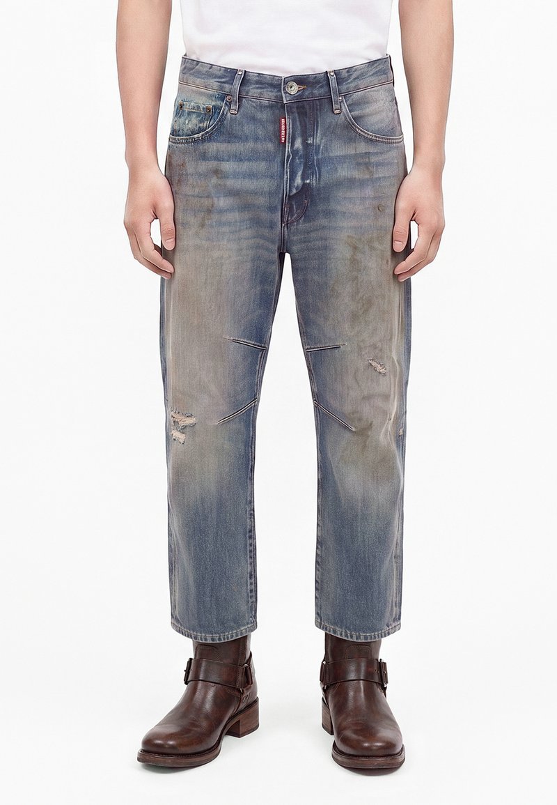 Homme portant un jean bleu délavé et usé avec des coutures aux genoux et des bottines en cuir marron, debout les mains détendues le long du corps.
