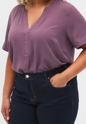 Frau trägt eine kurzärmelige lila V-Ausschnitt-Bluse, die in dunkelblaue, hoch taillierte Jeans gesteckt ist, mit einer Hand in der Hosentasche.