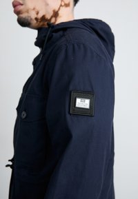 Navy blue hooded jacket with a buttoned chest pocket, adjustable drawstrings, and a square logo patch on the left sleeve.