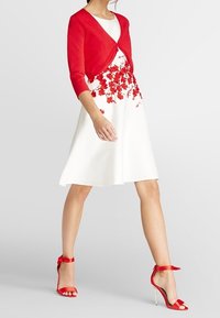 Rød strikket cardigan over en hvid kjole med rød blomsterbroderi. Sammen med røde sandaletter med remme. Glatte teksturer og tætsiddende silhuet.