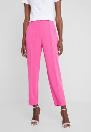 Adulto che indossa pantaloni dritti a vita alta di colore rosa acceso, camicia bianca a maniche corte e tacchi bianchi open toe con cinturino alla caviglia.
