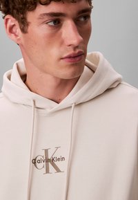 Młody mężczyzna z krótkimi kręconymi włosami, noszący kremową bluzę z kapturem z nadrukowanym z przodu logo Calvin Klein.