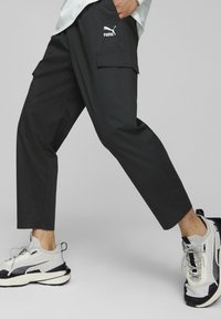 Pantalones cargo negros con un ajuste holgado, que cuentan con bolsillos laterales, detalles de costura y un logo blanco de Puma en el muslo. Combinados con zapatillas grises.