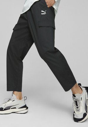 Pantalones cargo negros con un ajuste holgado, que cuentan con bolsillos laterales, detalles de costura y un logo blanco de Puma en el muslo. Combinados con zapatillas grises.
