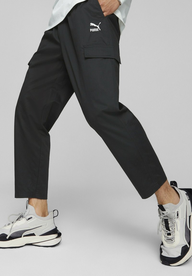 Pantalones cargo negros con un ajuste holgado, que cuentan con bolsillos laterales, detalles de costura y un logo blanco de Puma en el muslo. Combinados con zapatillas grises.