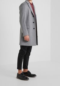 Abrigo de lana gris con un diseño a medida, cinco botones y bolsillos laterales, usado sobre una camisa burdeos y combinado con pantalones y zapatos negros.