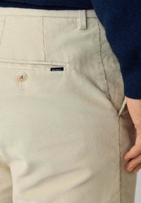 Pantalones de pana beige con un bolsillo trasero y detalle de botón. Cuenta con una pequeña etiqueta "Hackett" en azul marino. Tejido texturizado, suave y con un corte ajustado.