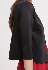 Zwarte cropped jas met 3/4 mouwen, gemaakt van soepele stof. Het ontwerp heeft een strakke silhouet, gecombineerd met een rode jurk eronder.