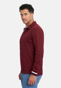 Camisa polo de manga larga en color burdeos con cuello, abertura de botones y detalles a rayas en los puños. Combinada con jeans azules, textura suave.