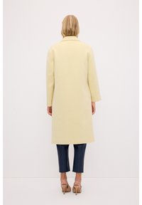 Abrigo largo amarillo de tela suave con un corte recto, con mangas dobladas y cuello con muesca, combinado con pantalones ajustados azul oscuro.